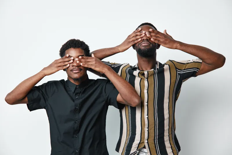 Two African men with dark skin covering their eyes with hands standing against white background. Casual clothing, friendship, gesture, and studio portrait of young adults.