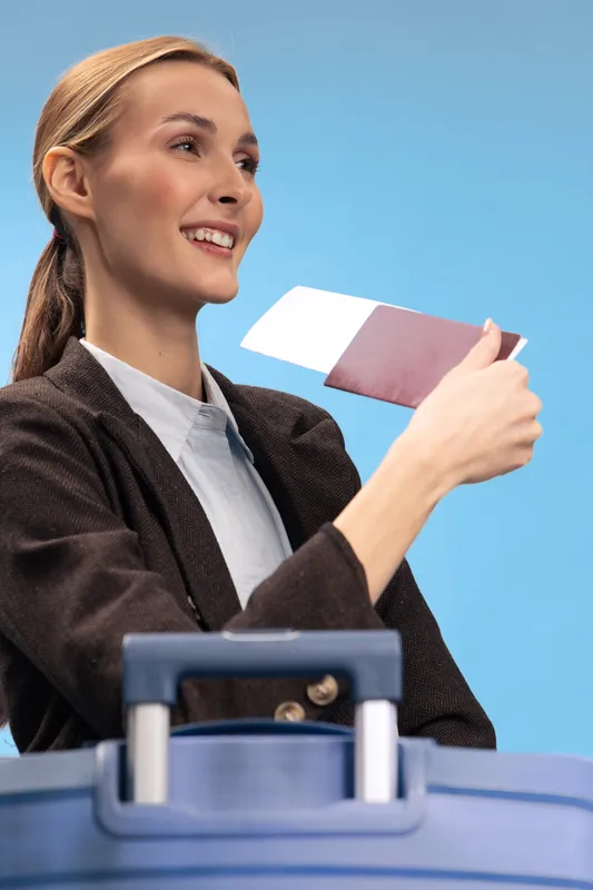Smiling woman holding passport and boarding pass near blue suitcase on light blue background. Travel concept with happy female traveler ready for vacation or business trip outdoors.