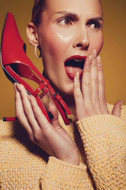 Surprised woman with short hair holds red high heel shoe near face, wearing yellow sweater and hoop earrings. Concept for fashion, style, and emotional expression in studio lighting.