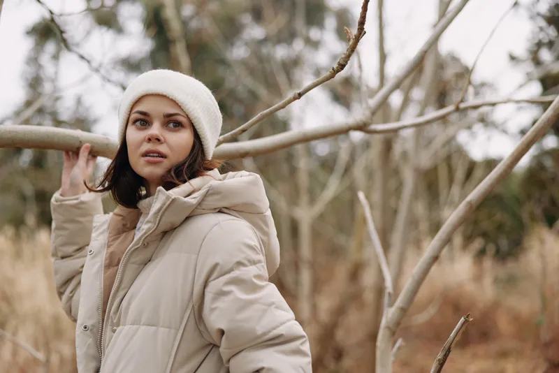Winter outdoor portrait of a woman in a beige padded coat and knitted beanie, standing beside leafless branches in a cold park, calm expression, soft natural light, fashion mood and seasonal setting