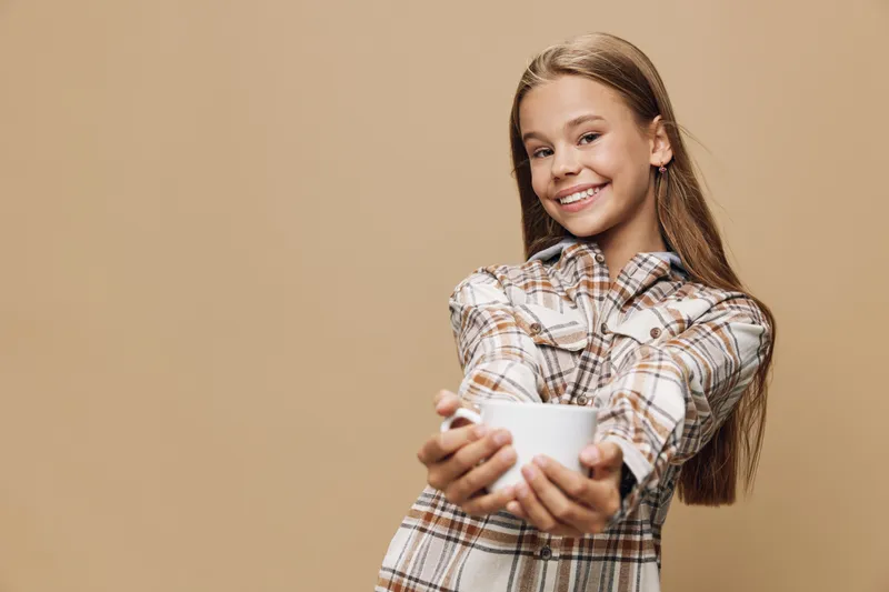Girl smiling and holding white cup with both hands wearing checkered shirt on beige background. Teenager happy and cheerful with long hair looking at camera and offering drink.