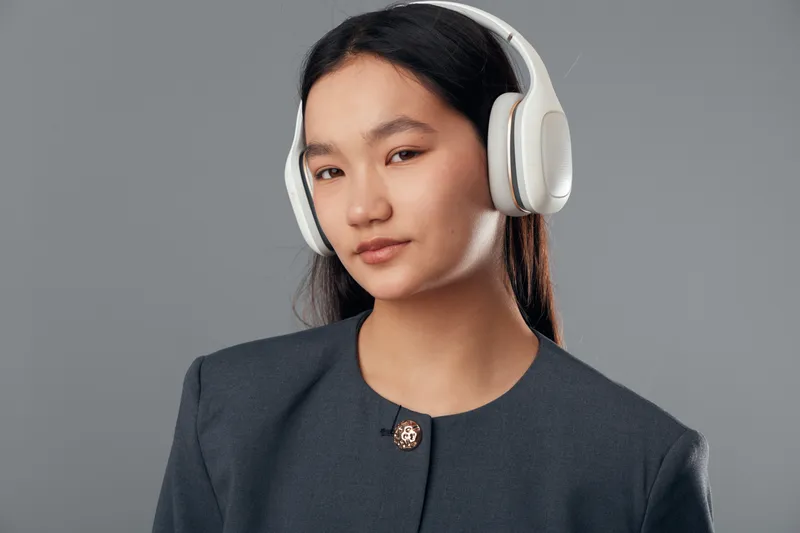 Young woman wearing white headphones and gray jacket posing with calm expression on gray background. Portrait of Asian female listening to music or audio content with long dark hair.