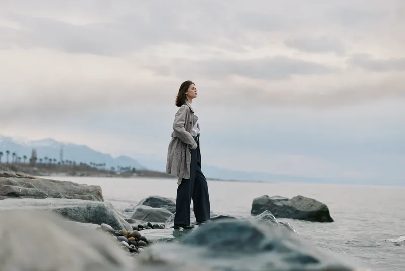 A contemplative woman in a long coat stands on rocky shore near calm water under cloudy sky. The peaceful scene shows natural landscape with distant mountains and soft light.