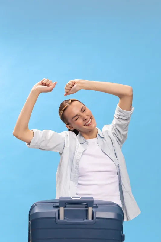 Young woman stretching arms happily behind a travel suitcase on blue background. Concept of vacation, travel preparation, relaxation, and positive emotions before trip.