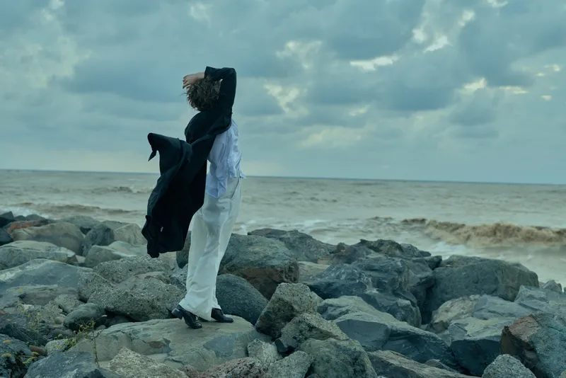 Fashion on rocky coast, windblown coat and white trousers by the sea, moody sky, dramatic silhouette as waves crash behind at dusk