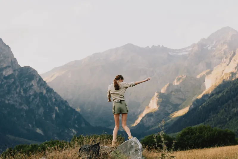Young woman stands on a sunlit rock amid rugged mountains, reaching toward the distant valley with a sense of adventure and freedom.
