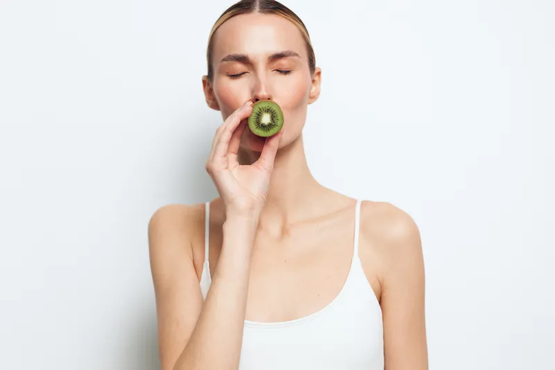 Woman holds kiwi fruit near lips, fresh healthy diet and nutrition concept, natural skincare and beauty routine, soft studio light on white background, eating for wellness today