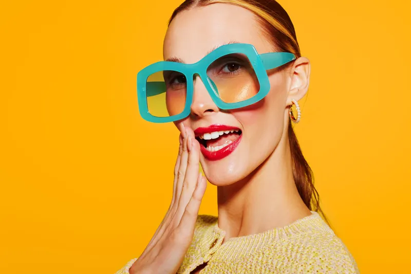 Smiling woman with large turquoise glasses and red lipstick poses against bright yellow background. She wears a light sweater and touches her cheek with a playful expression. Fashion concept.