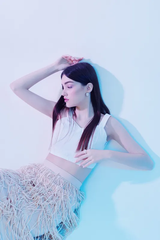 Young woman in a white crop top and textured skirt poses against a white wall with soft blue lighting. Concept for woman beauty fashion and modern style.