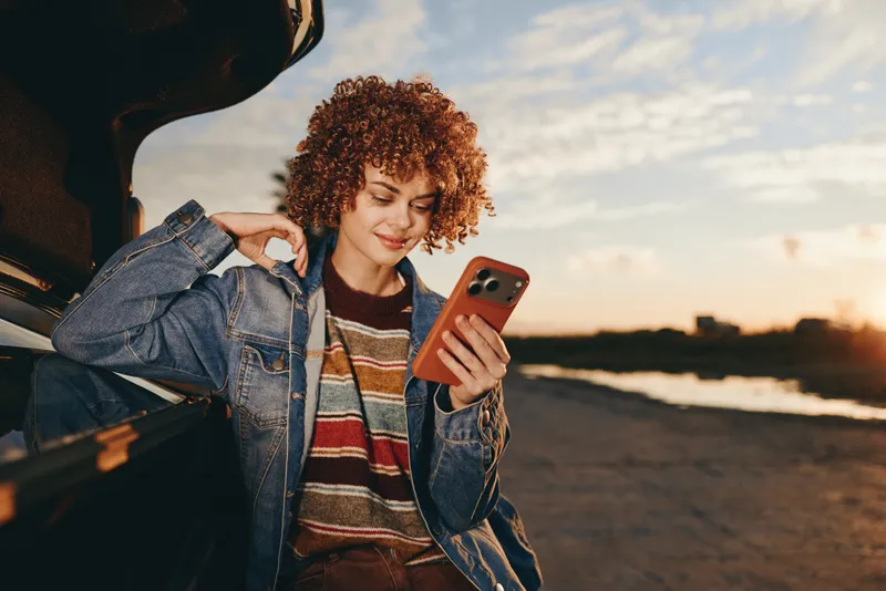 Woman lifestyle, curly haired woman in denim jacket checks smartphone near open car at sunset by the beach, holding credit card for online shopping, relaxed travel mood, warm light
