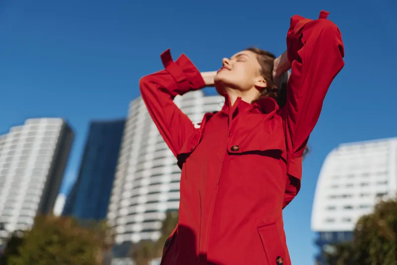 A young woman in a red coat stretches with closed eyes outdoors in an urban setting with modern white buildings and blue sky in the background, enjoying fresh air.