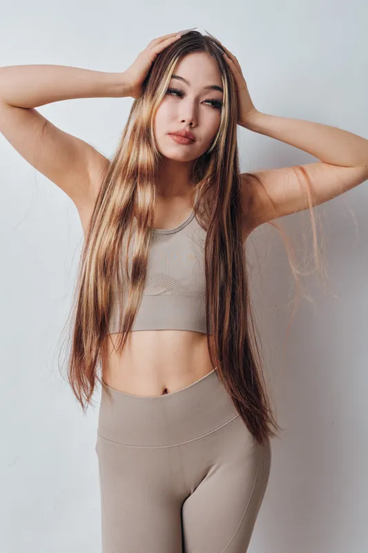 Young woman with long straight hair posing with hands on head wearing beige sportswear in studio. Casual outfit, natural look, fitness and beauty concept for healthy lifestyle and wellness.