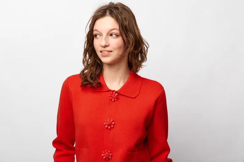 Young woman with curly brown hair wearing a red knitted sweater looks sideways with a slight smile. Studio portrait on white background with casual style and natural expression.