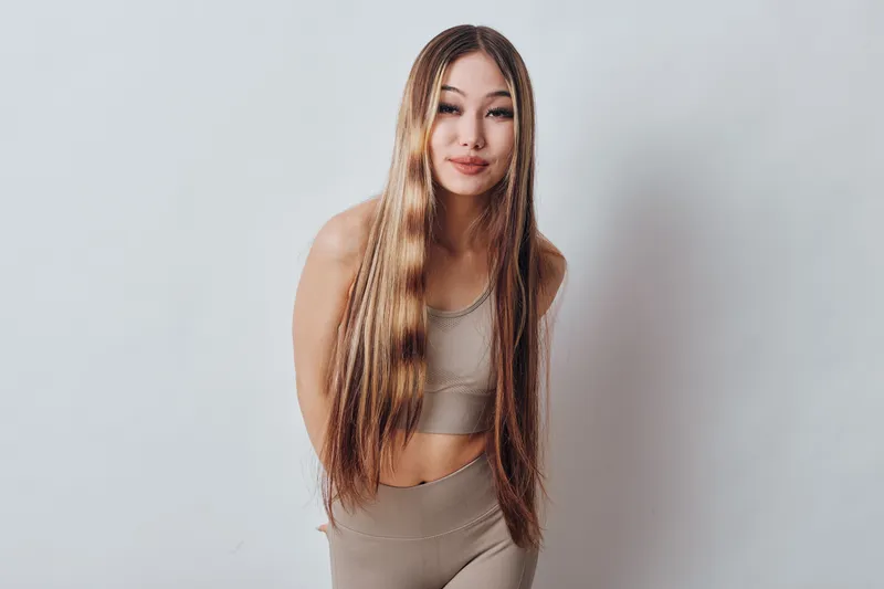 Young woman with long braided hair posing in neutral-colored sportswear standing against plain background. Fitness, beauty, lifestyle, health and confidence concept with casual style.