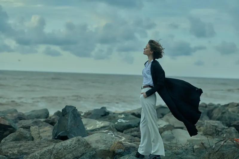 Woman stands on rocky shore, wind catching a long coat, ocean horizon behind, dramatic fashion scene by the sea, movement and strength in an outdoor coastal moment