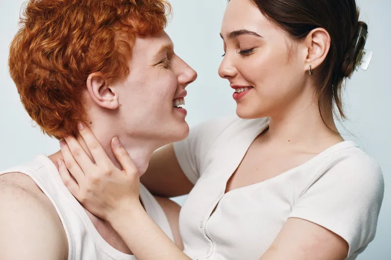 Young couple smiling and embracing close with affection and love in casual clothing against light background. Romantic relationship and happiness together.