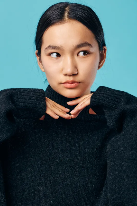 Young woman with dark hair wearing black sweater posing with hands under chin, looking sideways with thoughtful expression on blue background in studio portrait.