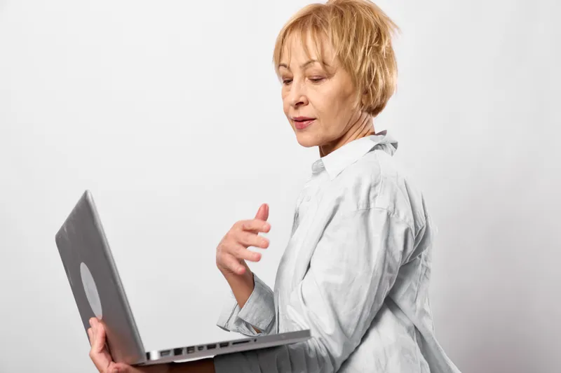 Senior woman with short blond hair holding a laptop indoors, side profile, concerned expression with hand gesture, minimal white background, soft daylight
