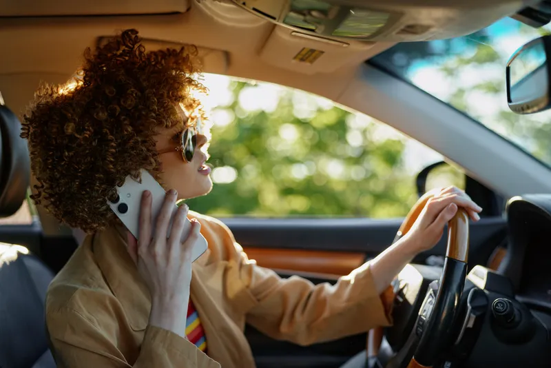 Curly haired woman driving a car while talking on a mobile phone. Beige jacket, warm sunlight through windows. Lifestyle shot highlighting independence, mobility, and everyday errands.