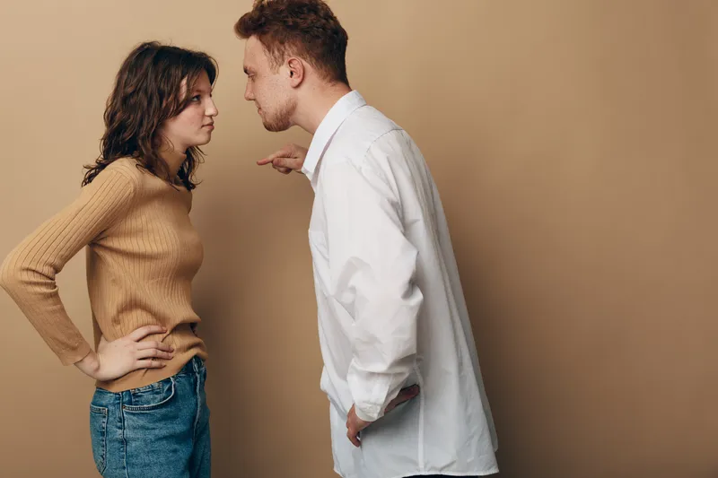 Angry young man in white shirt arguing with woman in brown sweater and jeans. Both show tense emotions and body language in a conflict situation on beige background.