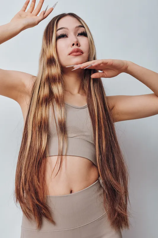 Woman with long hair posing with raised hands in studio. Female model wearing beige top and bottom against white background for fashion portrait.