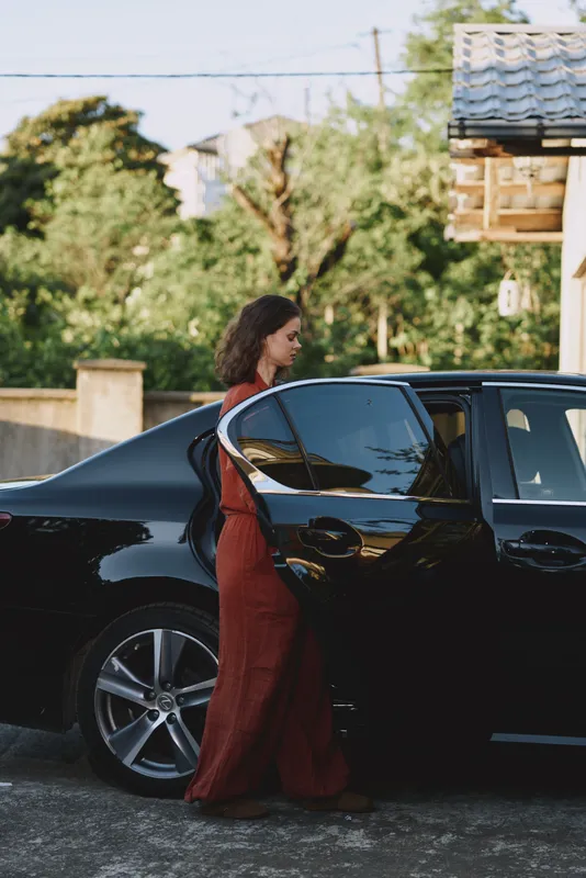 Woman in a flowing red dress approaches a sleek black car on a sunlit street, capturing a stylish moment of modern lifestyle, elegance, and confident arrival