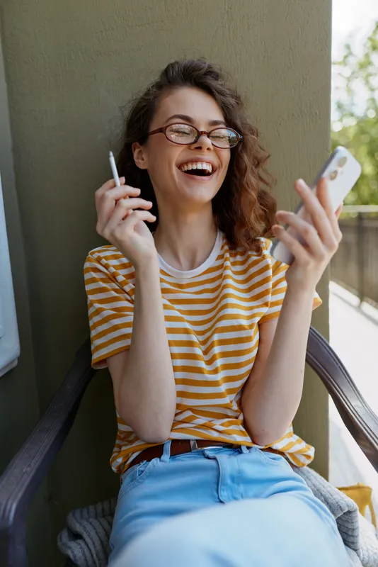 Casual woman enjoys a carefree moment on a balcony at home, smiling while talking on the phone and jotting notes, wearing a striped tee and light jeans.