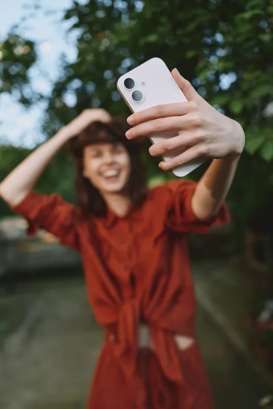 Young woman in a red dress takes a carefree selfie outdoors, smiling while extending her arm to capture the moment with a smartphone, her joyful pose conveying spontaneity and a relaxed lifestyle