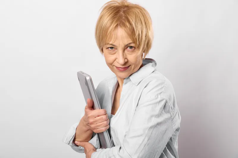 Woman senior holding smartphone, technology communication portrait indoors, white jacket, smiling, closeup studio lighting, neutral background, calm mood