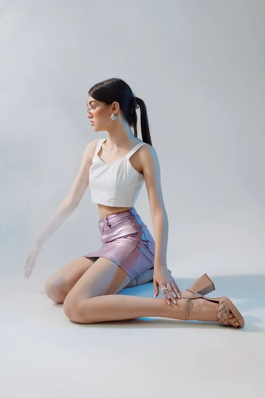 Young woman in a white crop top and metallic skirt kneels on the floor posing sideways. Studio shot with soft lighting. Concept of beauty fashion and modern style.