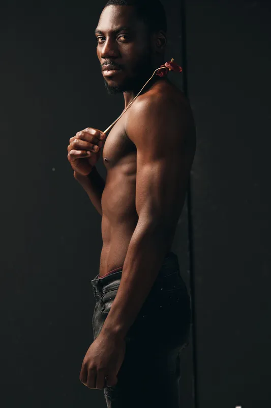 African man with muscular body and bare torso holding flower on shoulder in creative conceptual visual style. Dark skin, confident pose and artistic portrait with black background.