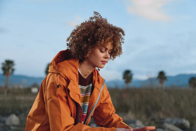 Woman with curly hair wearing orange jacket outdoors, relaxed lifestyle in nature, field and distant sky behind, warm sunlight, candid portrait with smile, under gentle wind, today