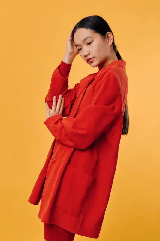 Woman in red oversized jacket with long dark hair standing on yellow background with thoughtful expression touching head with hand. Studio portrait of young female model in casual fashion pose.