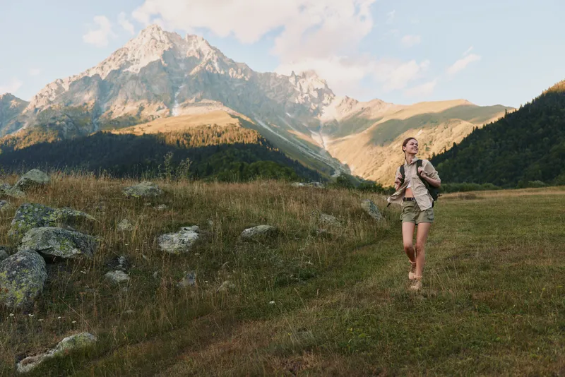 Woman hiker with backpack on a grassy trail, mountains in the background. Bright daylight, blue sky, alpine landscape, conveying freedom, vitality, and outdoor exploration.