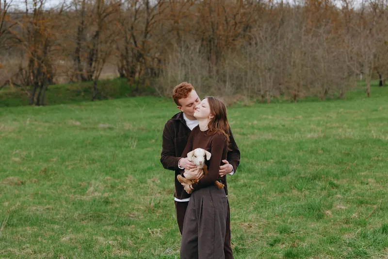 Young couple embraces in a green field while holding a small baby goat as they share a tender kiss during a calm outdoor lifestyle moment.