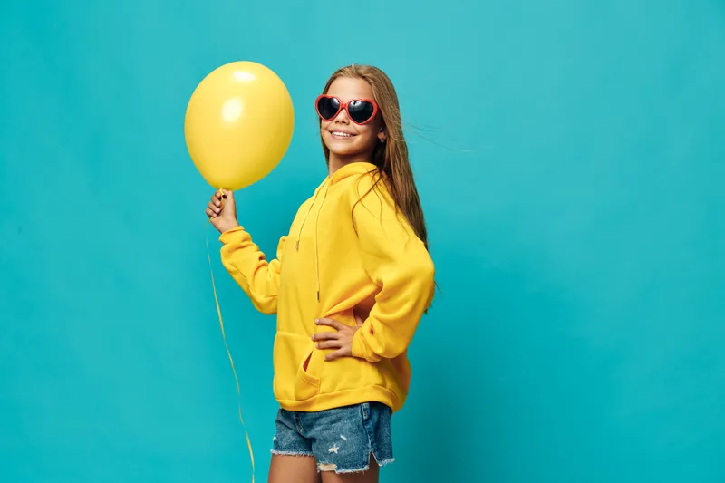 Young woman wearing sunglasses and yellow hoodie holding yellow balloon standing on blue background, smiling confidently with hand on hip and casual denim shorts.