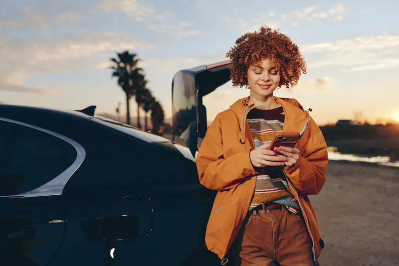 Woman lifestyle, a delivery courier in an orange jacket stands by a car at sunset, holding a smartphone and phone, with a backpack and warm evening light on the road.