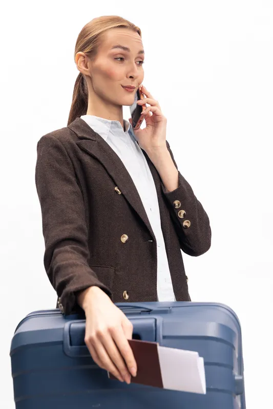 Young businesswoman in brown blazer holds blue suitcase and passport, talking on phone. Isolated on white background. Concept for travel, business trip, and communication.