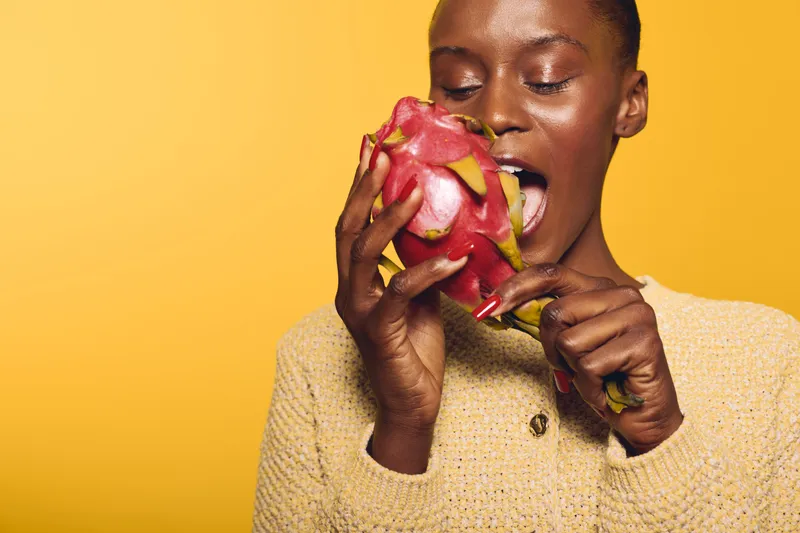 Young dark skinned African woman wearing a yellow sweater bites a fresh dragon fruit on yellow background. Concept of healthy eating, tropical fruit, natural food and lifestyle.