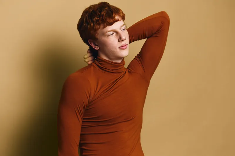Young man with red hair wearing brown turtleneck posing thoughtful with hand behind head against plain beige background in studio.