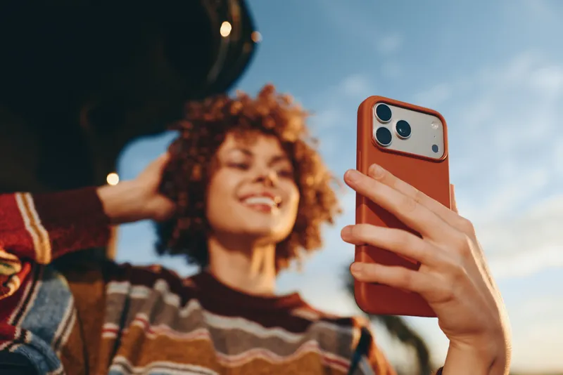 Woman selfie with phone, smiling curly hair, lifestyle outdoor moment, hand holds orange smartphone, blue sky background, closeup, striped sweater, cheerful relaxed mood