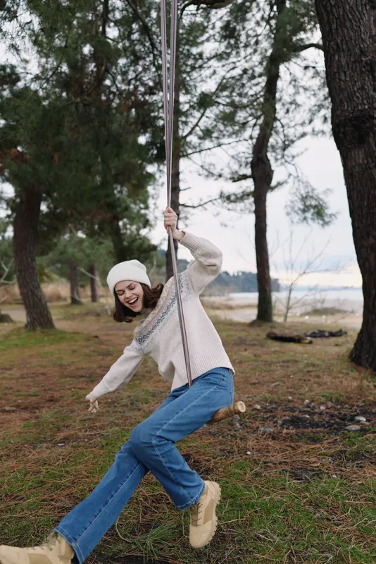 Outdoor swing moment with a girl in a knit sweater and jeans swinging beside trees by a lakeside, capturing playful movement and fresh nature.