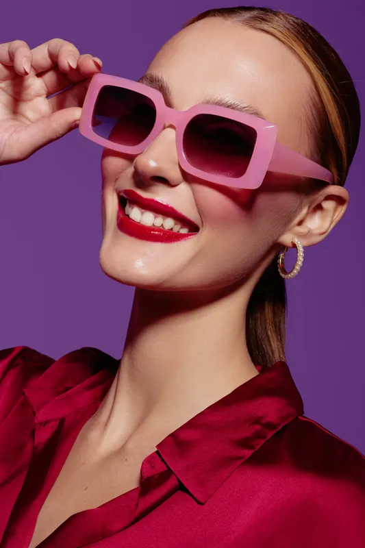 Smiling woman with pink sunglasses and red lipstick wears red satin blouse and gold hoop earrings on purple background. Concept of fashion, beauty, style, and confident mood.