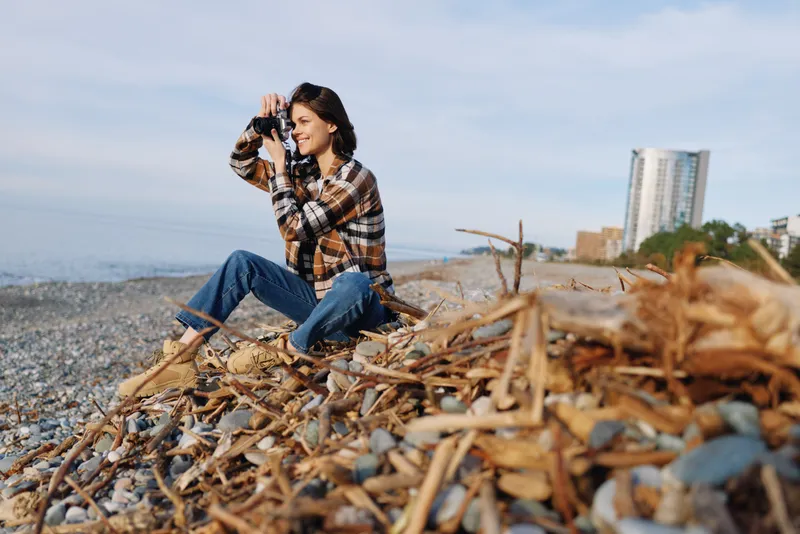 Woman photographer on a beach with camera, exploring a seascape and driftwood, wearing a plaid shirt and denim, focused mood under bright sky with calm waves and distant buildings in the background