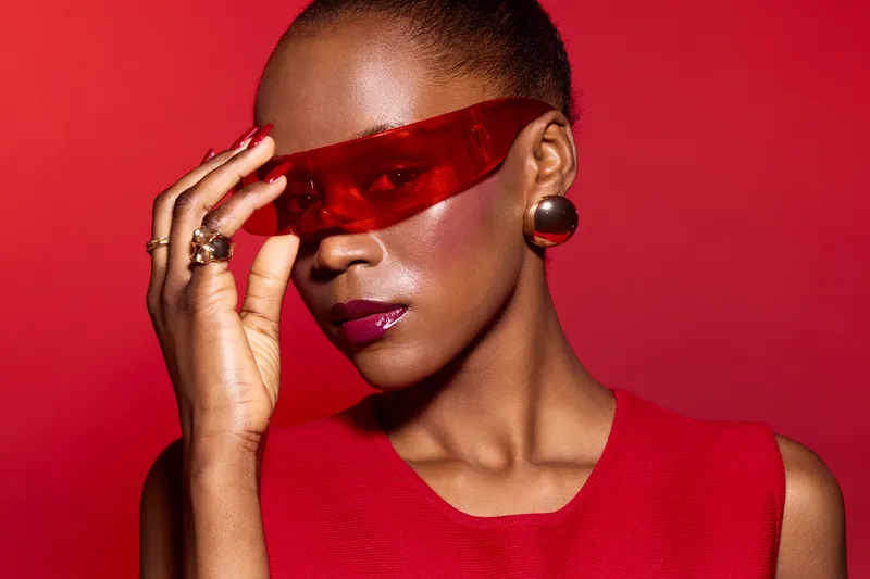 Stylish dark skinned African woman in red dress and red translucent glasses poses confidently on red background. Concept of fashion, elegance, beauty, and modern style.