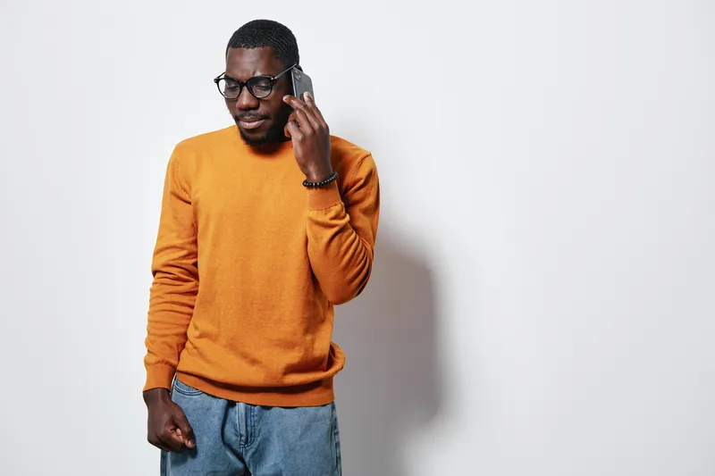 African American man with dark skin wearing glasses and orange sweater talking on smartphone against white background. Casual style, communication and technology concept with confident expression.