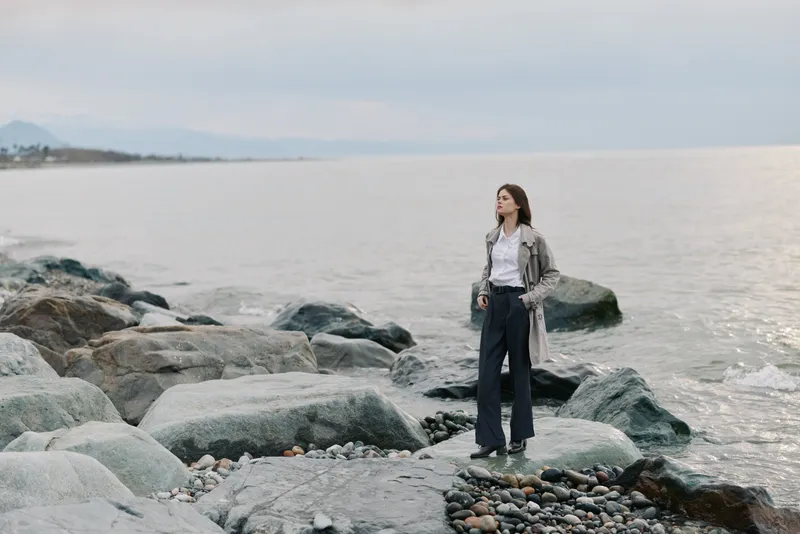 A thoughtful woman stands on rocky shore looking at the calm sea under an overcast sky. Casual outfit and serene mood convey contemplation and peaceful nature connection.
