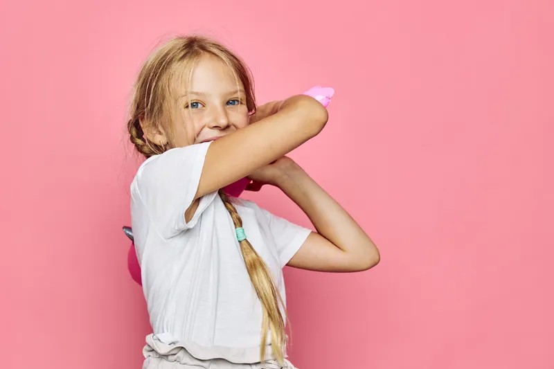 Happy young girl with blonde hair and braids wearing a white t-shirt poses playfully covering her face with her arm on a pink background. Joyful children concept.