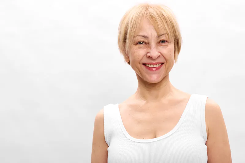 Smiling elderly woman portrait with short hair in a white sleeveless top on a clean white background. Calm confident mood, friendly senior lifestyle.