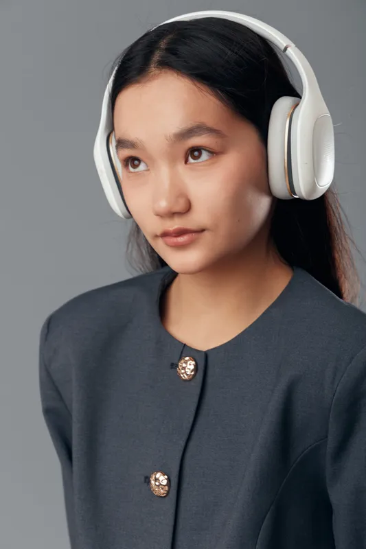 Young woman wearing wireless headphones and dark gray jacket listening to music or podcast with focused expression on gray studio background in closeup portrait view.